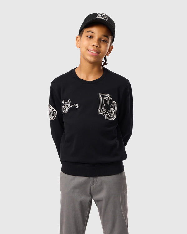 A person wearing the Psycho Bunny KIDS ZACK VARSITY SWEATSHIRT - B0S880D200, in black with chenille embroidery, along with a matching black cap, stands against a plain background. The sweatshirt showcases writing on the chest and left sleeve, as well as a vintage-inspired logo. The person is smiling with their hands behind their back.