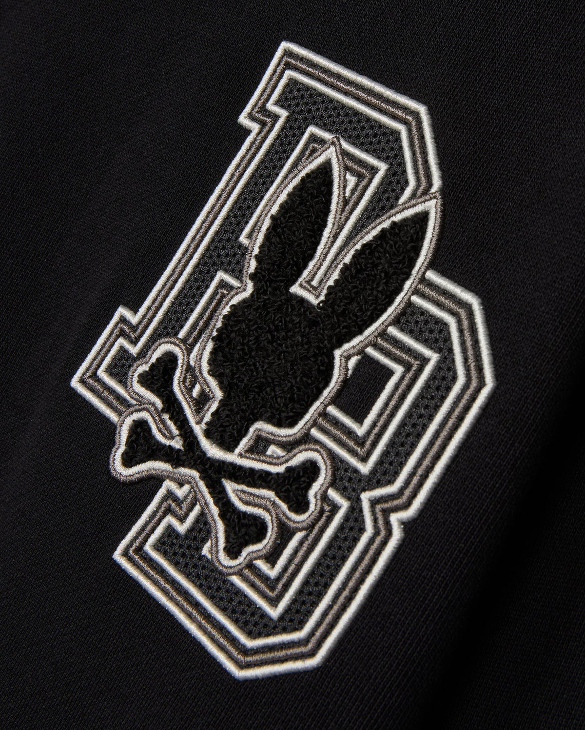 The KIDS ZACK VARSITY SWEATSHIRT by Psycho Bunny features a striking chenille embroidery design with a large letter 'B' outlined in white. Overlapping it is a playful skull and crossbones motif, where a bunny head sits above crossed bones, all in black and white outlines, reminiscent of a vintage-inspired logo.