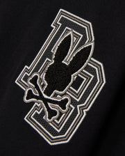 The KIDS ZACK VARSITY SWEATSHIRT by Psycho Bunny features a striking chenille embroidery design with a large letter 'B' outlined in white. Overlapping it is a playful skull and crossbones motif, where a bunny head sits above crossed bones, all in black and white outlines, reminiscent of a vintage-inspired logo.
