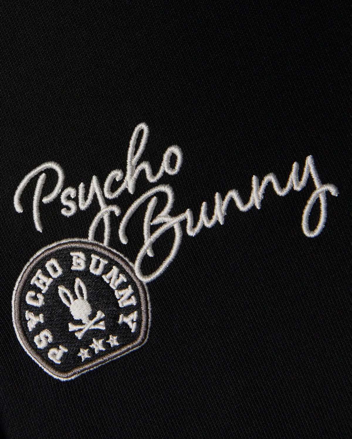 Adorning the black fabric of the KIDS ZACK VARSITY SWEATSHIRT - B0S880D200 by Psycho Bunny, white chenille embroidery spells out "Psycho Bunny." Below it, a vintage-inspired logo displays a circular emblem that surrounds a bunny head with crossbones, evoking the look of a classic varsity sweatshirt.