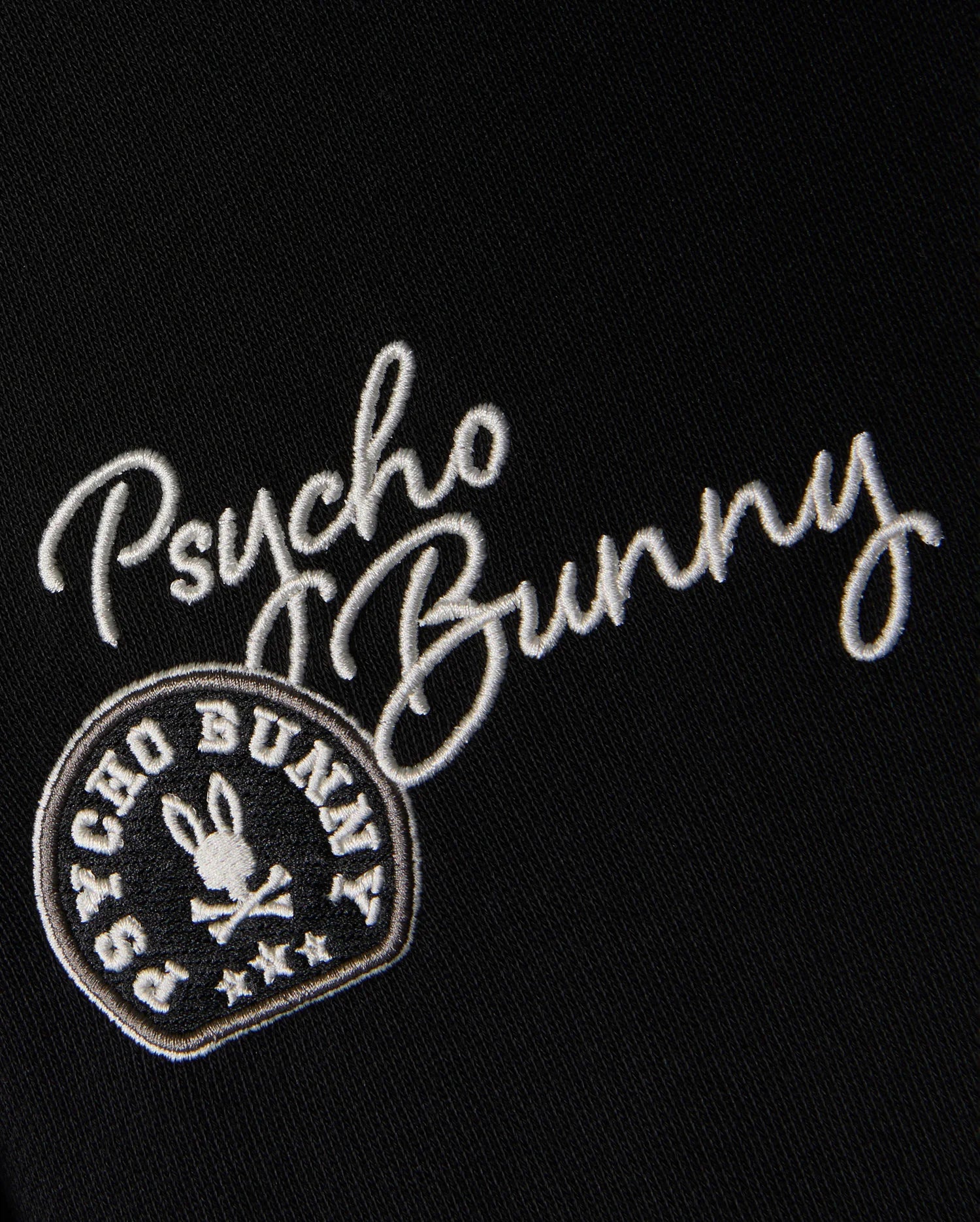 Adorning the black fabric of the KIDS ZACK VARSITY SWEATSHIRT - B0S880D200 by Psycho Bunny, white chenille embroidery spells out "Psycho Bunny." Below it, a vintage-inspired logo displays a circular emblem that surrounds a bunny head with crossbones, evoking the look of a classic varsity sweatshirt.