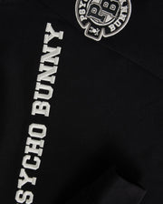 Close-up of the Kids Zack Varsity Sweatshirt by Psycho Bunny in black, featuring white "PSYCHO BUNNY" text on the sleeve and a vintage-inspired logo with chenille embroidery letters "PB" on the upper chest area.