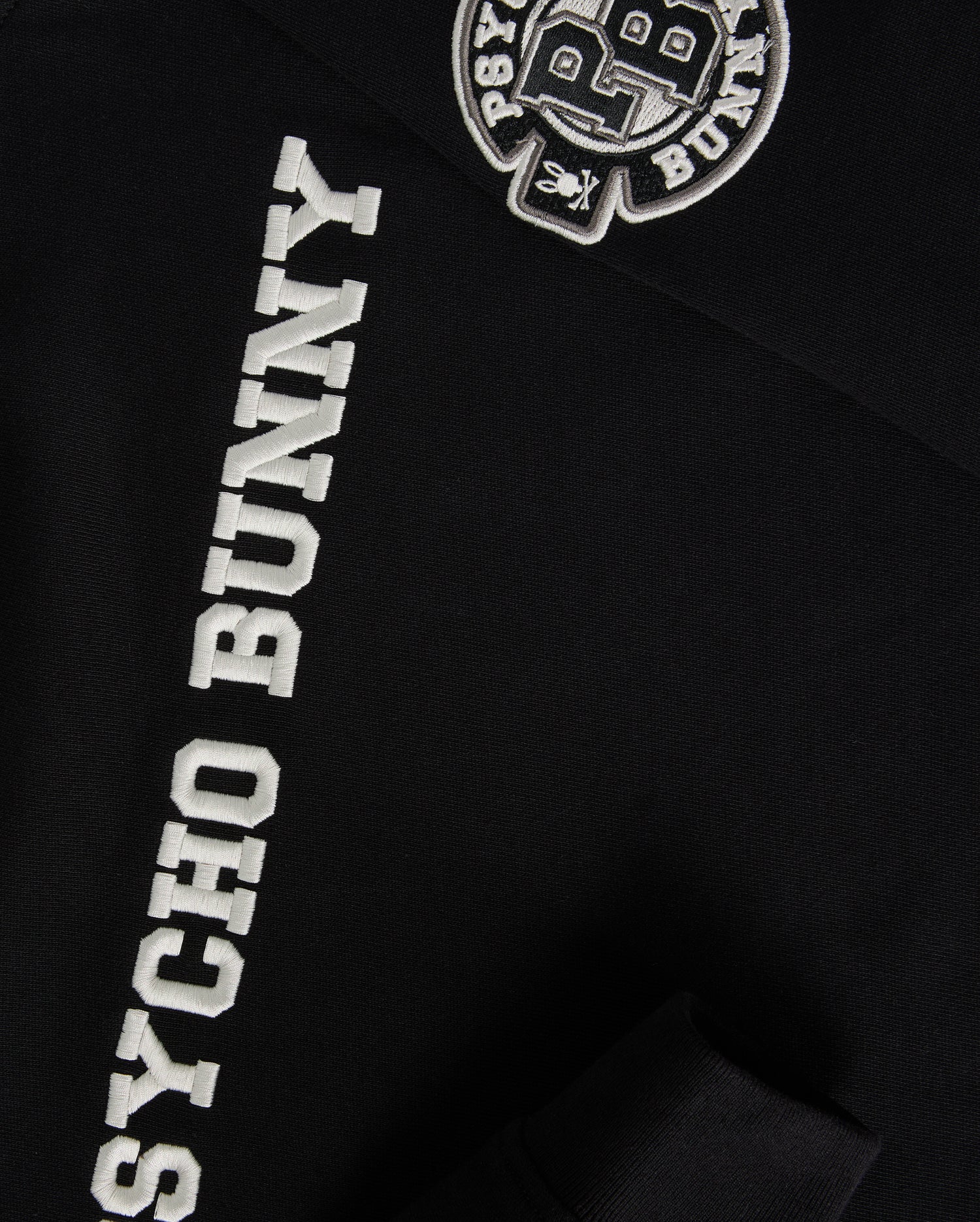 Close-up of the Kids Zack Varsity Sweatshirt by Psycho Bunny in black, featuring white "PSYCHO BUNNY" text on the sleeve and a vintage-inspired logo with chenille embroidery letters "PB" on the upper chest area.