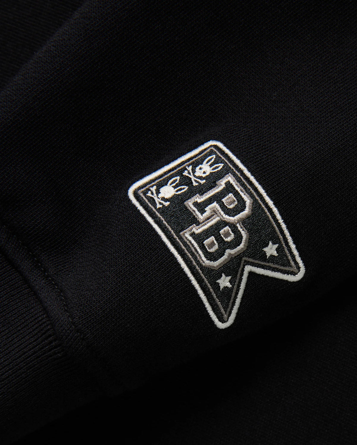 Close-up of the black fabric on the KIDS ZACK VARSITY SWEATSHIRT - B0S880D200 by Psycho Bunny, highlighting a vintage-inspired logo with chenille embroidery. The embroidered patch displays two bees above the letters "PB" and two stars below, all in white thread for a striking contrast against the dark background.