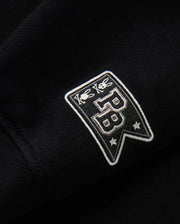 Close-up of the black fabric on the KIDS ZACK VARSITY SWEATSHIRT - B0S880D200 by Psycho Bunny, highlighting a vintage-inspired logo with chenille embroidery. The embroidered patch displays two bees above the letters "PB" and two stars below, all in white thread for a striking contrast against the dark background.