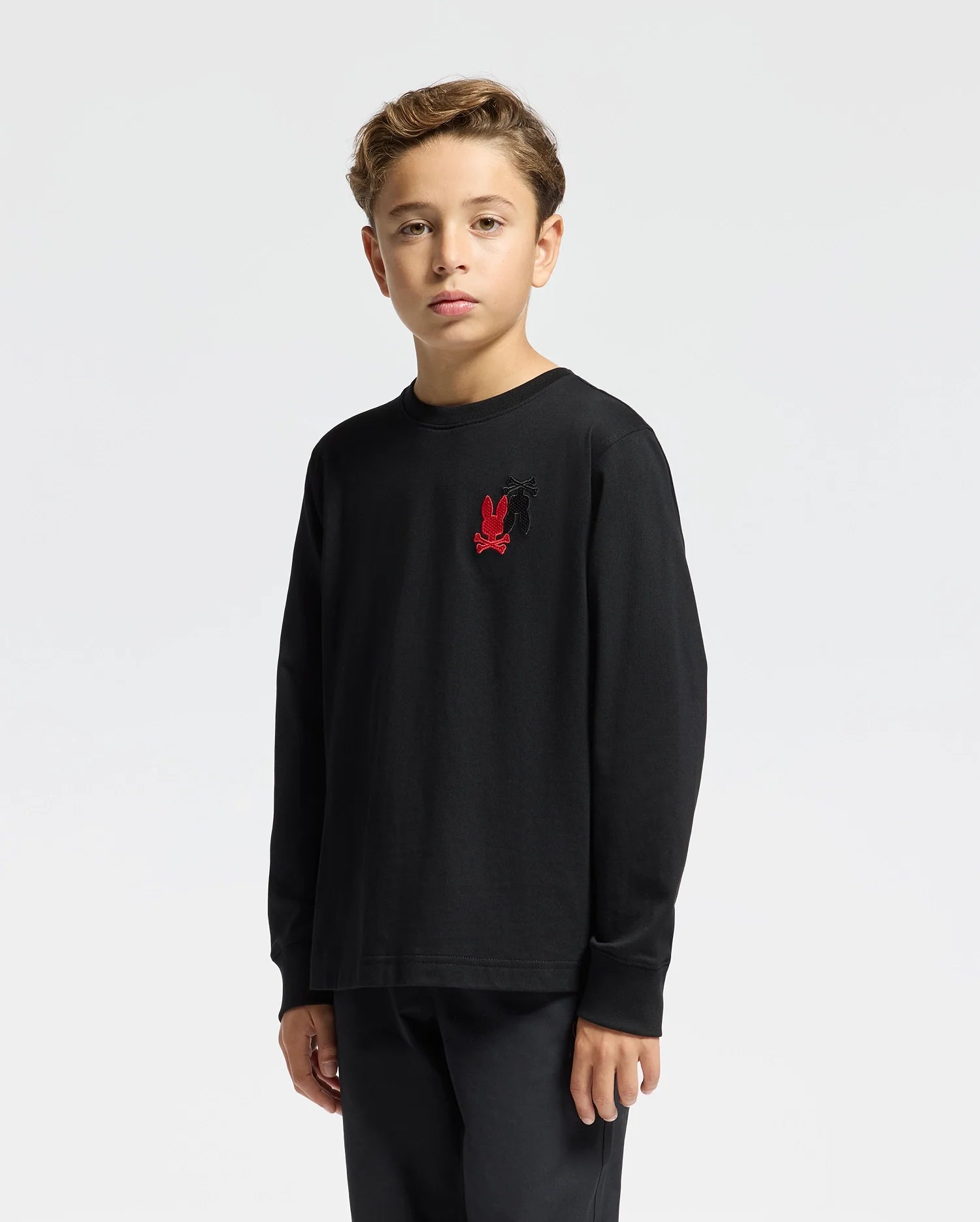 A young boy wearing a black Psycho Bunny Kids Pierce Long Sleeve Back Graphic Tee, featuring a striking red graphic on the front, stands against a plain white background.