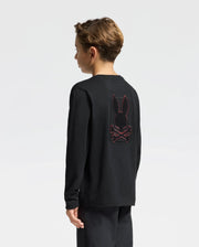 A young boy stands in profile, facing left, dressed in a Psycho Bunny KIDS PIERCE LONG SLEEVE BACK GRAPHIC TEE - B0T178E200, crafted from soft Pima Cotton. The shirt features an embroidered bunny and crossbones graphic on the back. The background is a plain, light gray.
