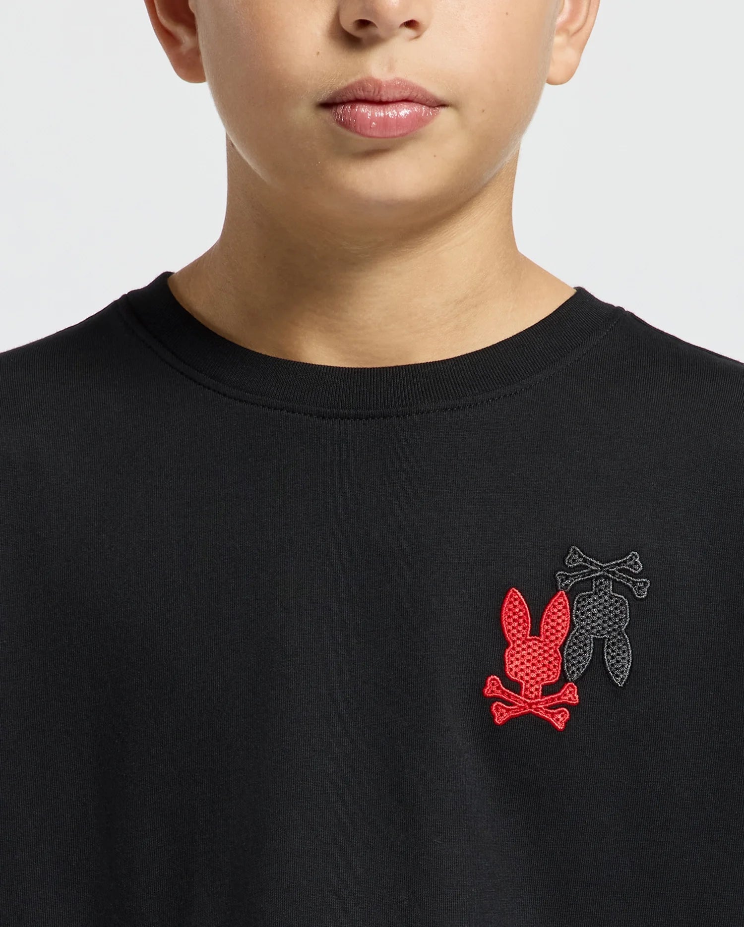 A person wearing the Kids Pierce Long Sleeve Back Graphic Tee by Psycho Bunny, with embroidered designs over the chest depicting two stylized rabbit heads—one in red and the other in black—each accompanied by crossed bones underneath, set against a plain, light-colored background.