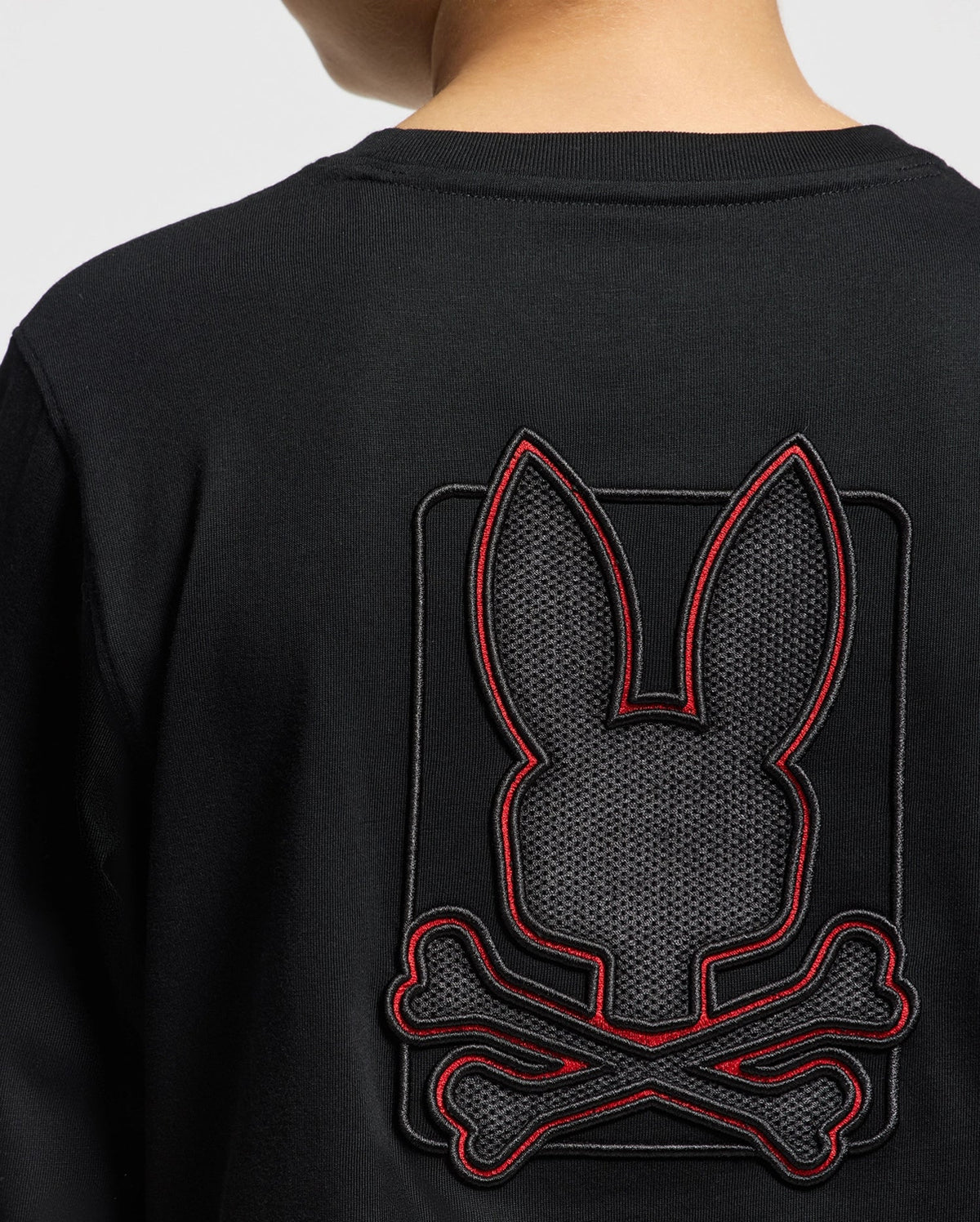 Someone is wearing a black Psycho Bunny Kids' Pierce Long Sleeve Back Graphic Tee, which showcases an embroidered bunny design featuring a stylized bunny head with long ears above crossed bones, all outlined with striking red stitching.