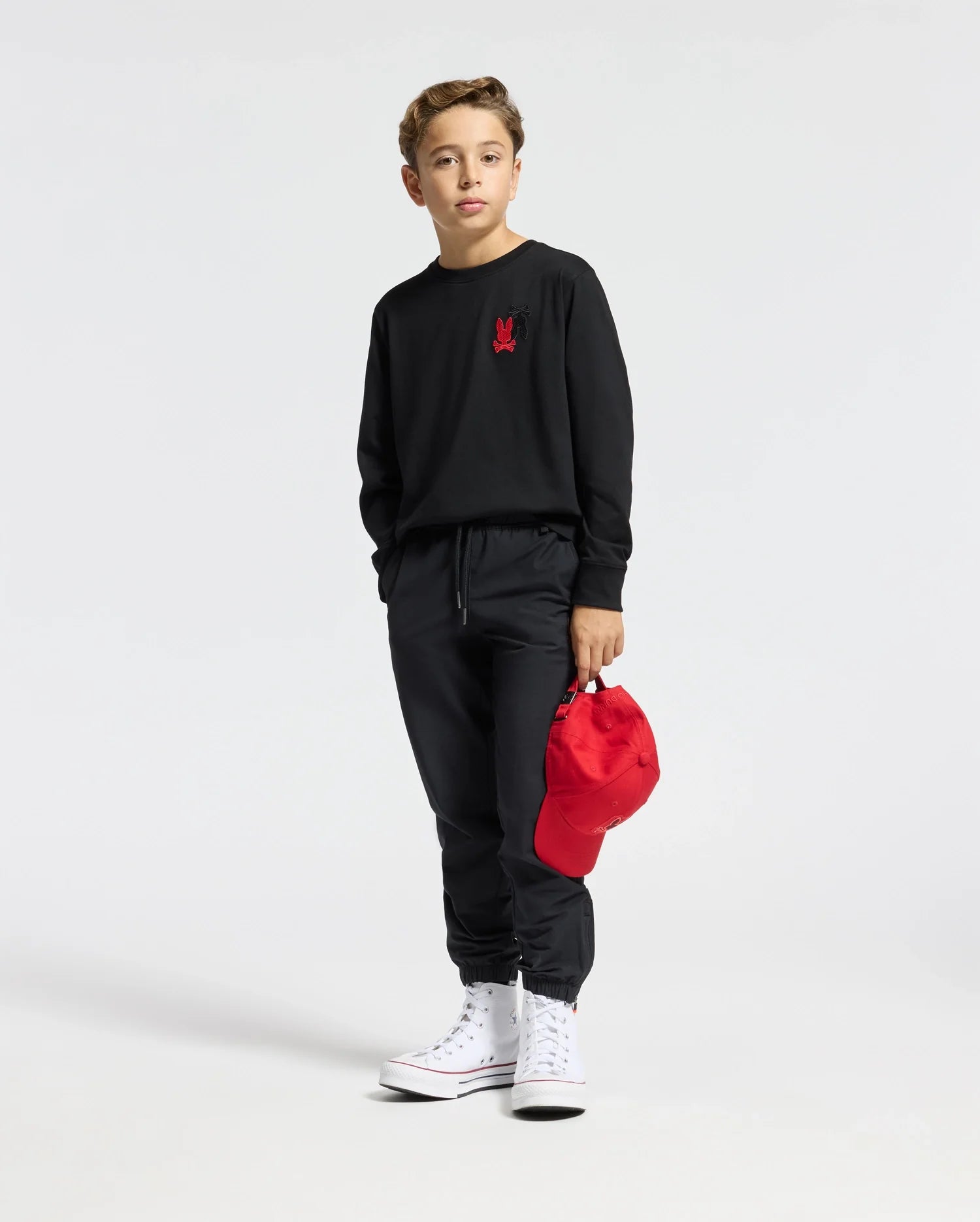 A young boy stands dressed in a black Psycho Bunny KIDS PIERCE LONG SLEEVE BACK GRAPHIC TEE paired with pants and white high-top sneakers. He holds a red baseball cap in his left hand. The shirt, crafted from soft pima cotton, is adorned with a small red logo on the chest. The background is plain white.