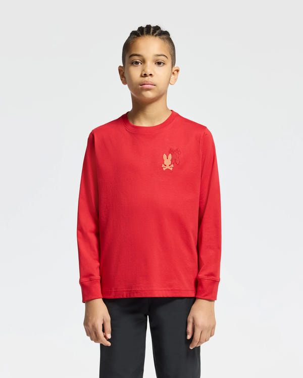 A young individual with braided hair stands against a plain background, wearing a red KIDS PIERCE LONG SLEEVE BACK GRAPHIC TEE by Psycho Bunny in soft Pima cotton, adorned with an embroidered bunny on the chest, paired with black pants. They have a neutral expression.