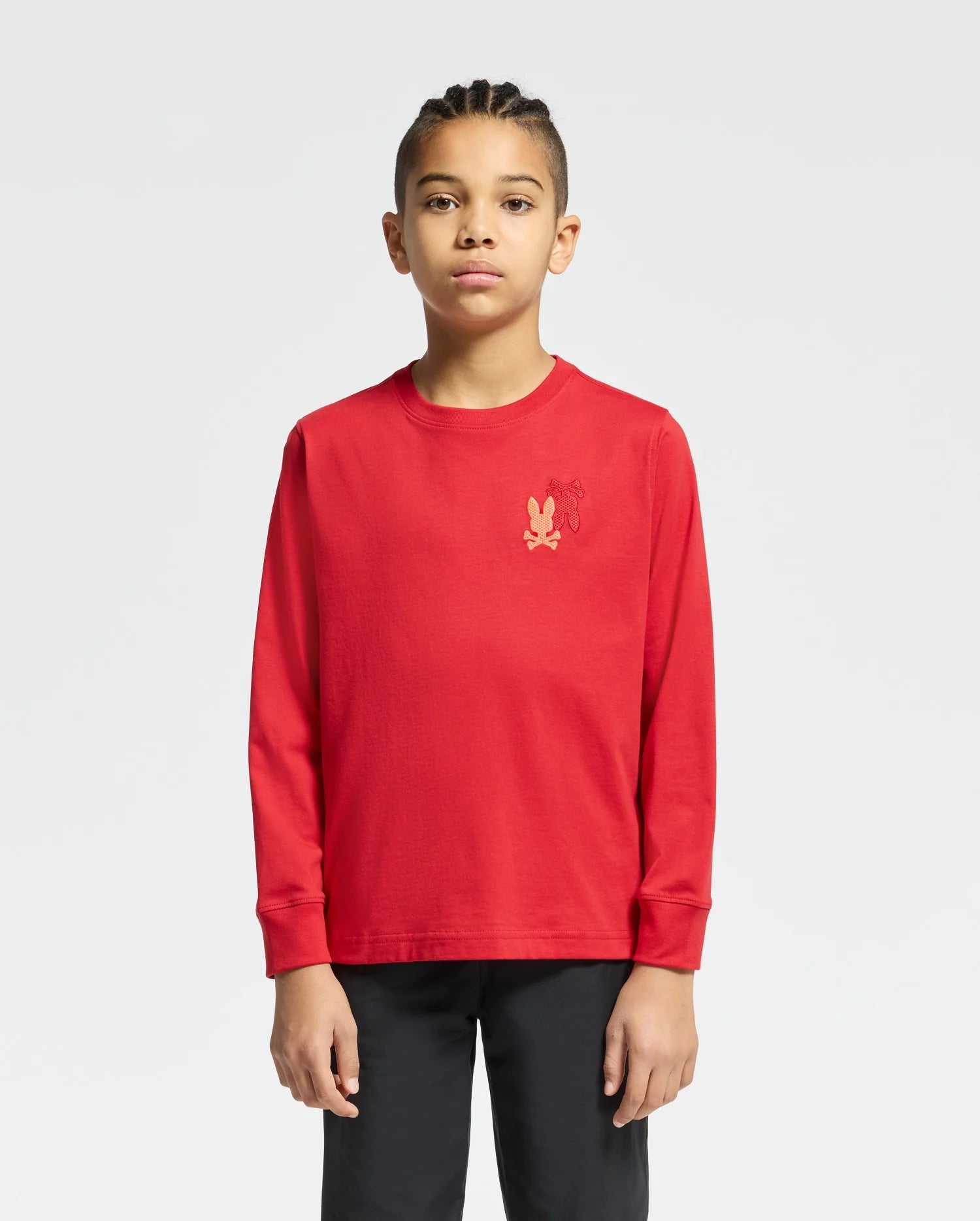 A young individual with braided hair stands against a plain background, wearing a red KIDS PIERCE LONG SLEEVE BACK GRAPHIC TEE by Psycho Bunny in soft Pima cotton, adorned with an embroidered bunny on the chest, paired with black pants. They have a neutral expression.