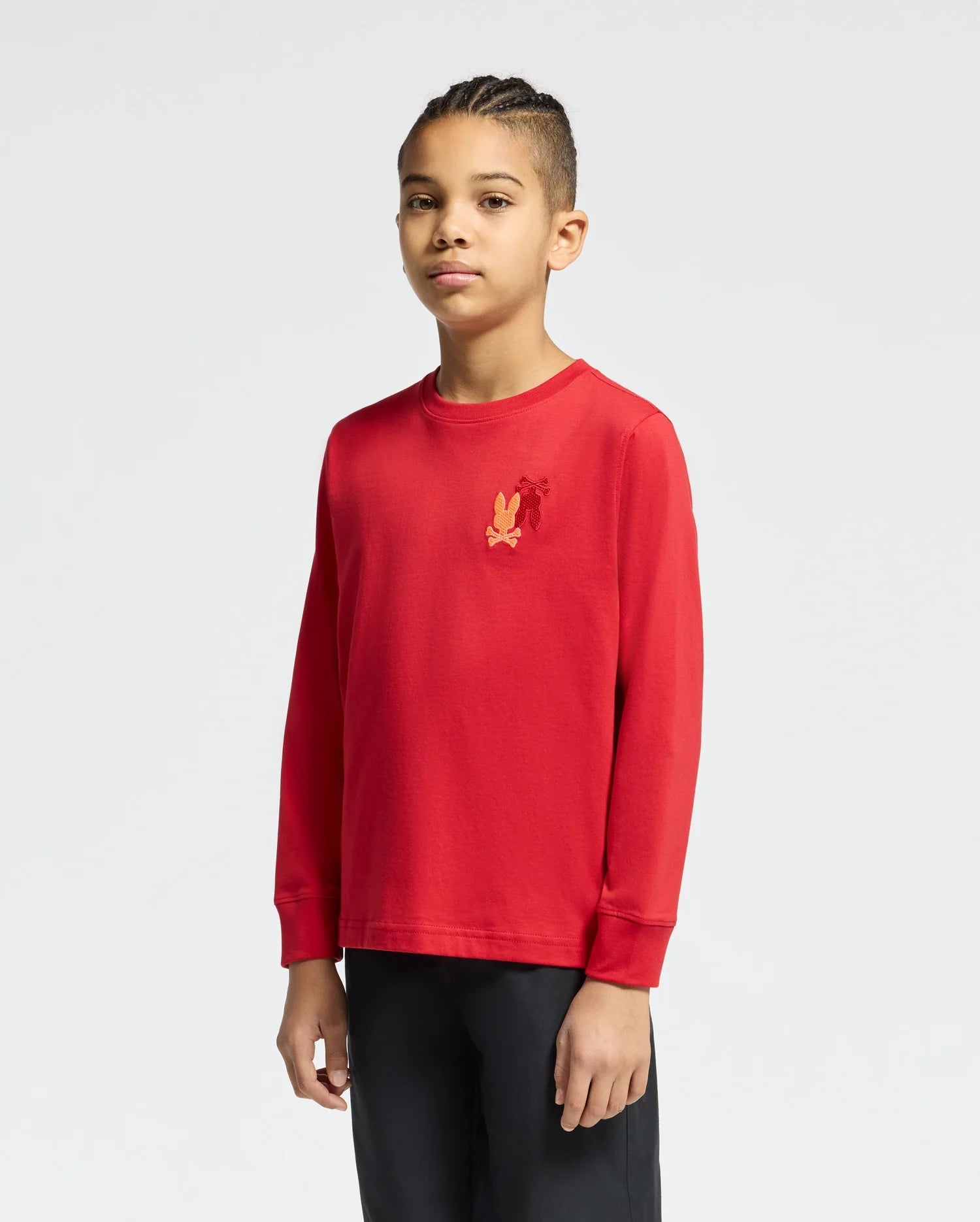 A young person stands facing forward against a plain background, wearing a red Psycho Bunny Kids' Pierce Long Sleeve Back Graphic Tee, featuring an embroidered bunny on the chest, and paired with black pants.