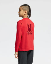A young individual with braided hair wears the Psycho Bunny KIDS PIERCE LONG SLEEVE BACK GRAPHIC TEE in a vibrant red, featuring an embroidered bunny and crossbones design on the back. Made from luxurious pima cotton, they pose sideways against a plain white background, allowing the design to truly stand out.
