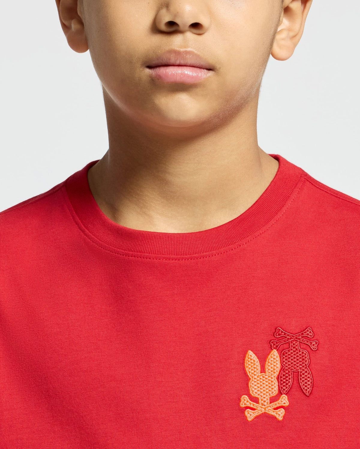 The image features a person wearing the "KIDS PIERCE LONG SLEEVE BACK GRAPHIC TEE - B0T178E200" by Psycho Bunny, which displays embroidered orange and red leaf designs and crossed bone shapes on the chest. The vibrant top, probably crafted from luxurious Pima cotton, is highlighted in the photo to focus on the lower face and upper torso.