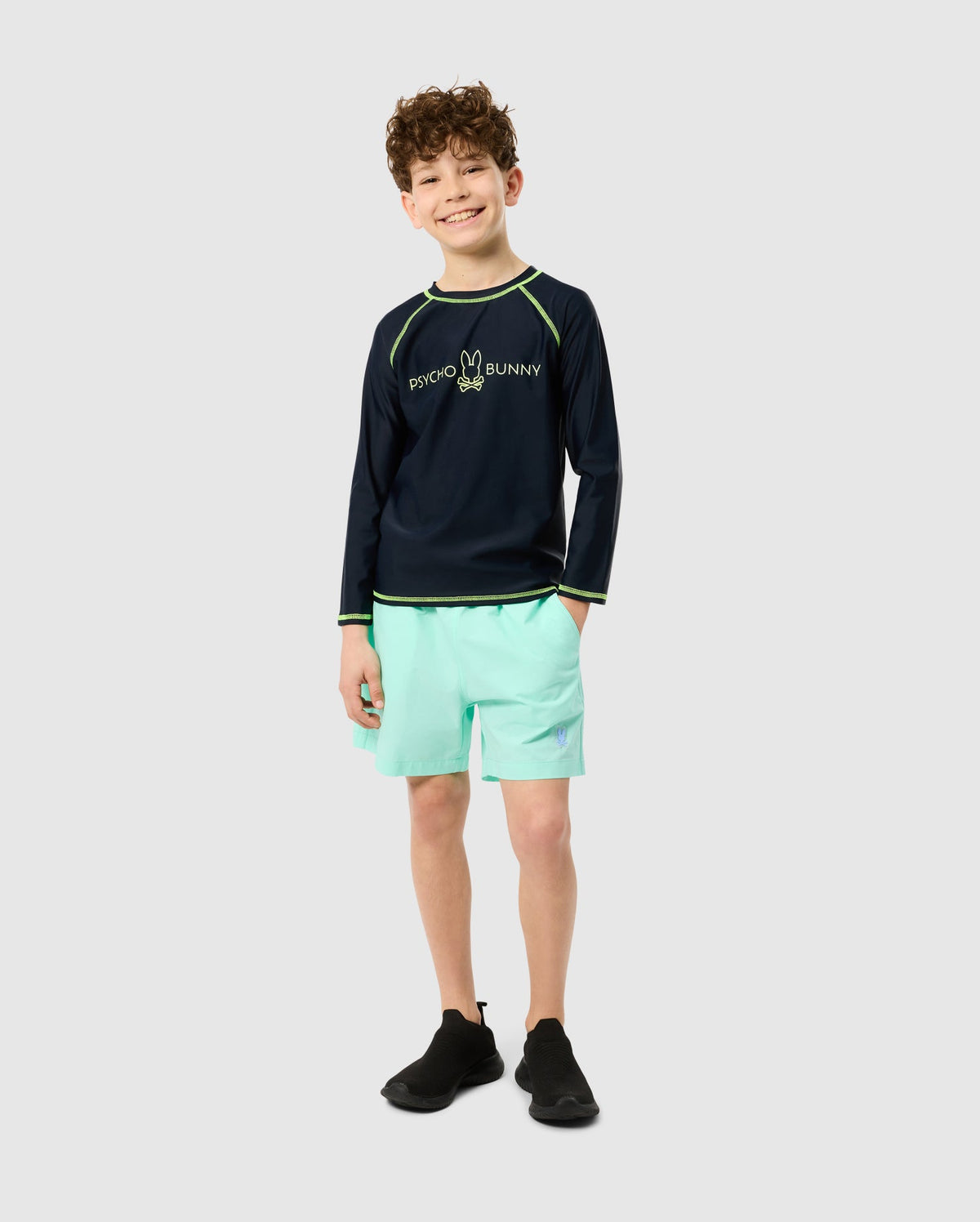 A smiling child with curly hair wears Psycho Bunny's KIDS LONG SLEEVE RASH GUARD - B0T557C200, offering UPF 50+ protection. Paired with light turquoise shorts and quick-drying black shoes, they stand against a plain light gray background.