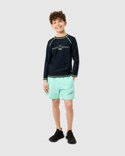 A smiling child with curly hair wears Psycho Bunny's KIDS LONG SLEEVE RASH GUARD - B0T557C200, offering UPF 50+ protection. Paired with light turquoise shorts and quick-drying black shoes, they stand against a plain light gray background.