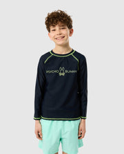 A curly-haired boy smiles in a navy KIDS LONG SLEEVE RASH GUARD (B0T557C200) by Psycho Bunny, designed for quick-drying comfort. Paired with light green shorts, the outfit is ideal for fun under the sun against a neutral light gray backdrop that highlights his cheerful style.