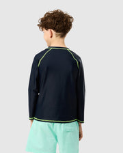 A curly-haired child, facing away from the camera, wears Psycho Bunny's KIDS LONG SLEEVE RASH GUARD - B0T557C200 in navy with green stitching and light turquoise shorts. The outfit provides UPF 50+ protection against a plain gray backdrop.