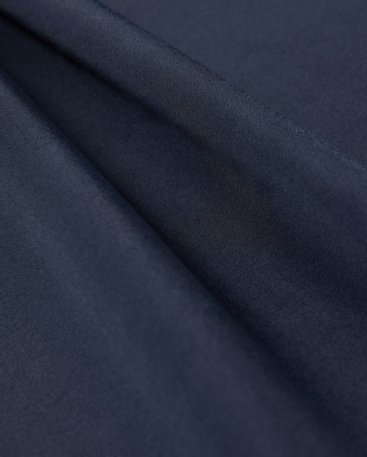 Close-up of folded, dark navy blue fabric from the Psycho Bunny KIDS LONG SLEEVE RASH GUARD - B0T557C200, showcasing its smooth texture and slight sheen. This fabric is ideal for a kids' rash guard with UPF 50+ protection, layered to create gentle folds and shadows.