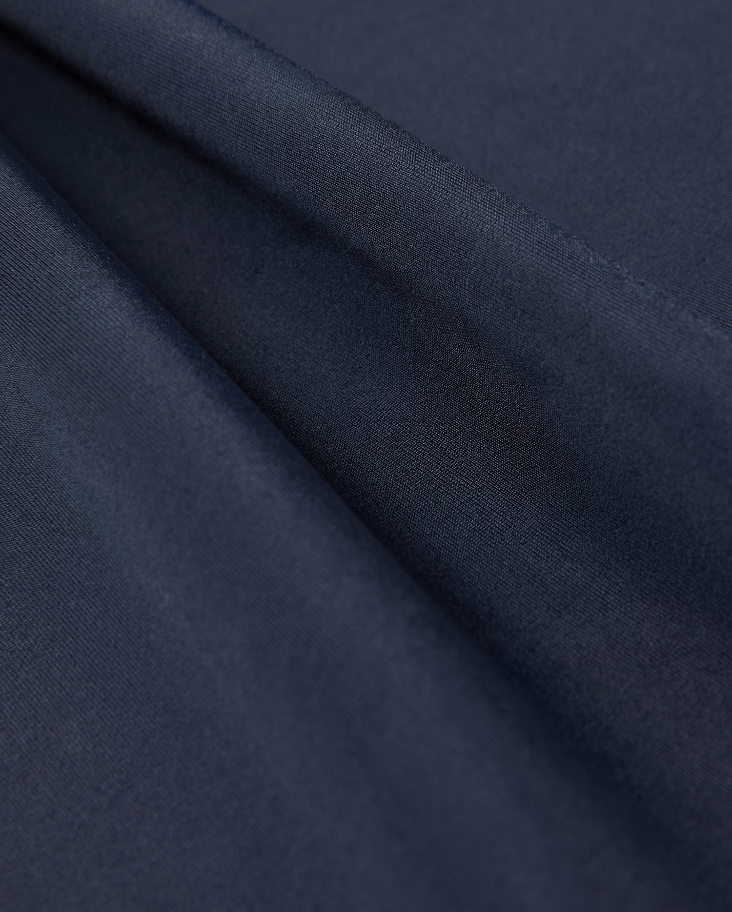Close-up of folded, dark navy blue fabric from the Psycho Bunny KIDS LONG SLEEVE RASH GUARD - B0T557C200, showcasing its smooth texture and slight sheen. This fabric is ideal for a kids' rash guard with UPF 50+ protection, layered to create gentle folds and shadows.