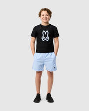 A cheerful young boy wearing a Psycho Bunny black short sleeve KIDS SAN LEON ANTI-UV RASHGUARD - B0T645C200 with a white rabbit design, light blue shorts, and black sneakers, standing against a plain white background.
