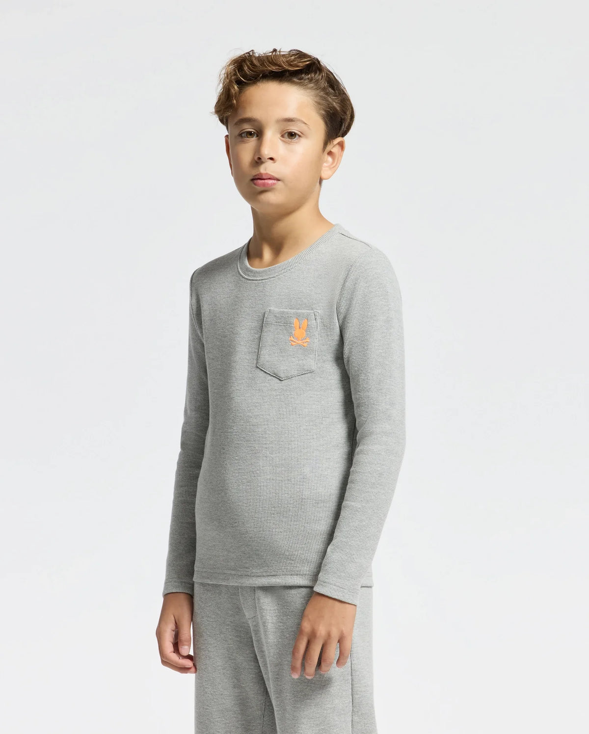A young boy stands against a plain white background, wearing a light gray Kids Val Waffle Long Sleeve Tee from Psycho Bunny in soft Pima cotton, which features an embroidered orange emblem on the chest pocket. He pairs it with matching gray pants. His expression is neutral and his hair is short and styled.