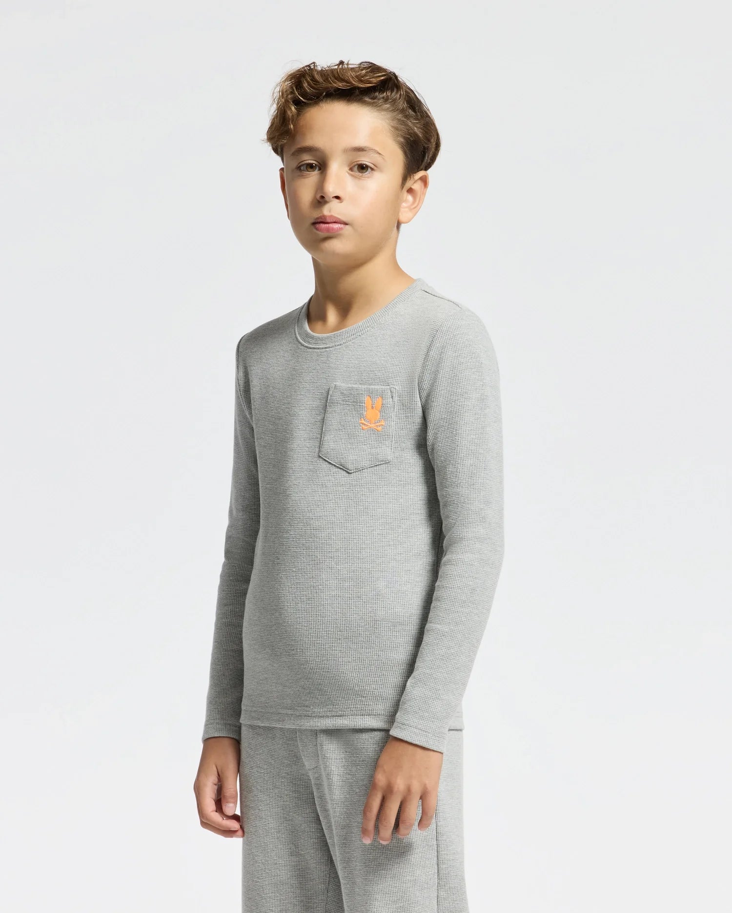 A young boy stands against a plain white background, wearing a light gray Kids Val Waffle Long Sleeve Tee from Psycho Bunny in soft Pima cotton, which features an embroidered orange emblem on the chest pocket. He pairs it with matching gray pants. His expression is neutral and his hair is short and styled.