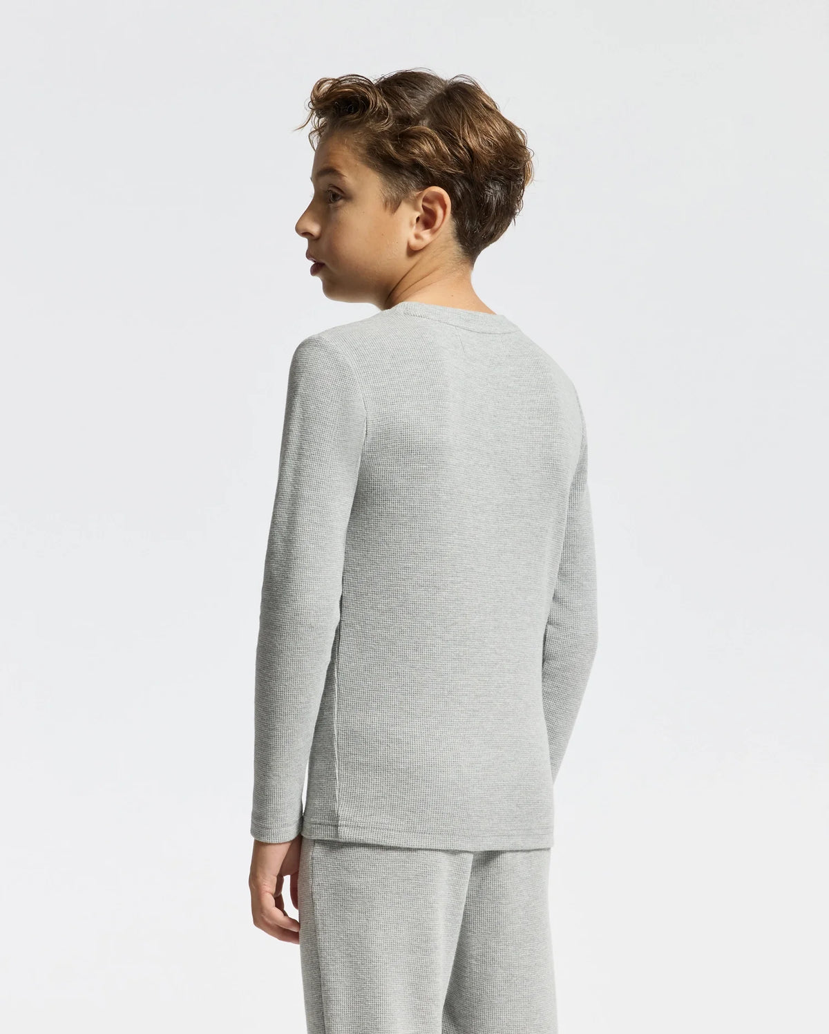 A young person with short curly hair is sporting an embroidered light gray Kids Val Waffle Long Sleeve Tee by Psycho Bunny, along with matching pants. They are shown from the side and back against a plain white background.
