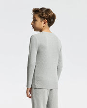 A young person with short curly hair is sporting an embroidered light gray Kids Val Waffle Long Sleeve Tee by Psycho Bunny, along with matching pants. They are shown from the side and back against a plain white background.