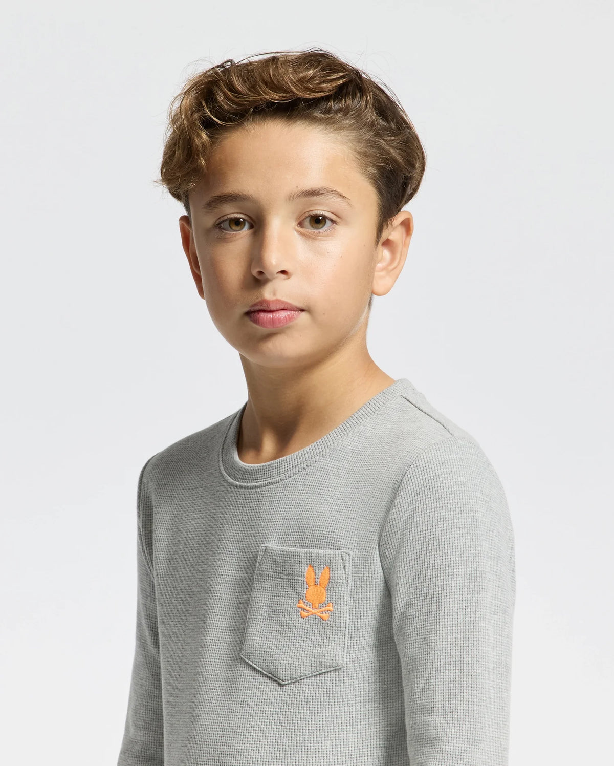 A young boy with brown hair stands against a plain white background, wearing a light gray Psycho Bunny KIDS VAL WAFFLE LONG SLEEVE TEE. Made from soft Pima Cotton, this shirt features a small embroidered orange bunny logo on the chest pocket. He looks directly at the camera with a neutral expression.