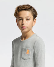 A young boy with brown hair stands against a plain white background, wearing a light gray Psycho Bunny KIDS VAL WAFFLE LONG SLEEVE TEE. Made from soft Pima Cotton, this shirt features a small embroidered orange bunny logo on the chest pocket. He looks directly at the camera with a neutral expression.