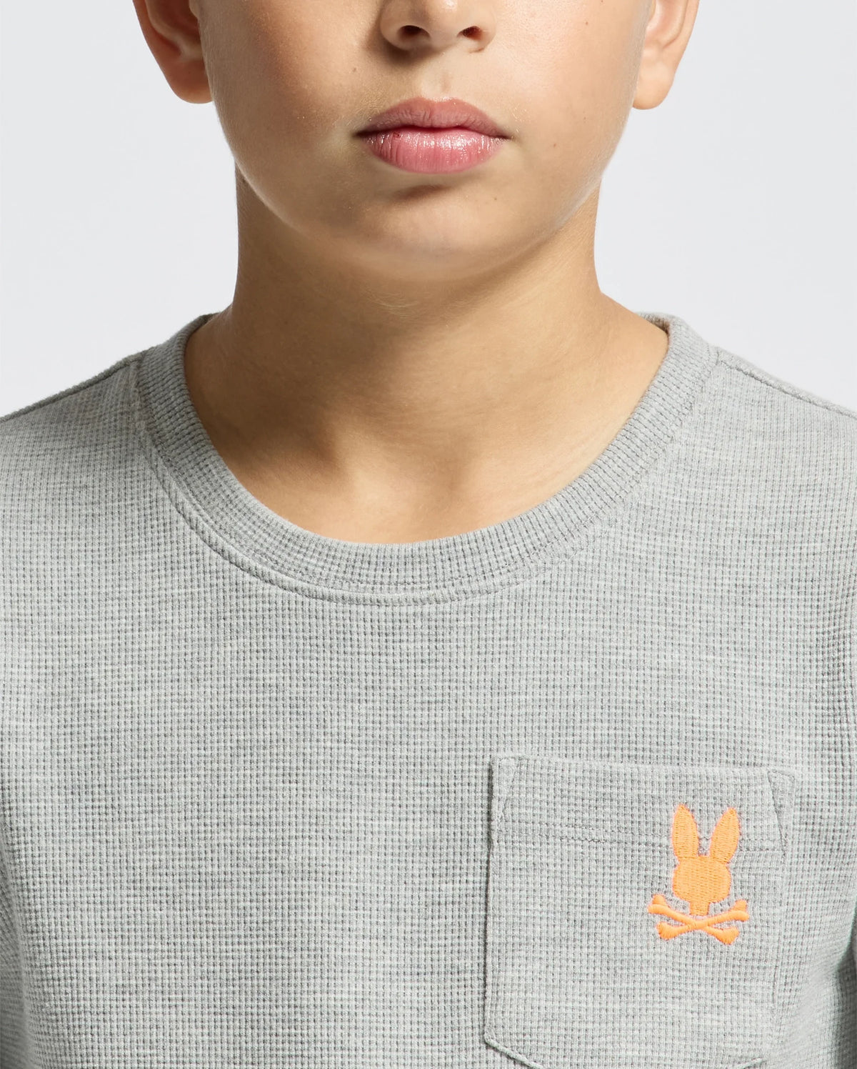 A person wearing a gray textured KIDS VAL WAFFLE LONG SLEEVE TEE (Product Code: B0T672E200) by Psycho Bunny, featuring an orange embroidered bunny logo on the chest pocket. The background is plain white.