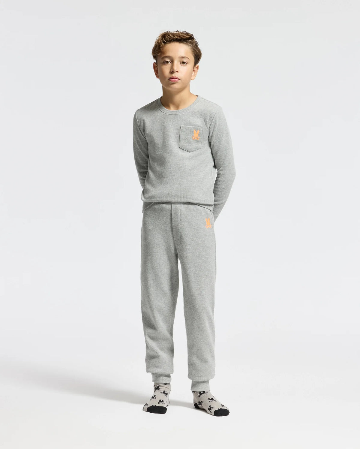 A young boy stands wearing the Psycho Bunny KIDS VAL WAFFLE LONG SLEEVE TEE, which comes in a gray color with a matching top and bottom set. The pajamas feature an embroidered orange emblem on both the chest and pants. He is also wearing socks with a black and white pattern against a plain white background.