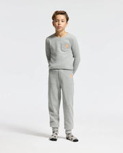 A young boy stands wearing the Psycho Bunny KIDS VAL WAFFLE LONG SLEEVE TEE, which comes in a gray color with a matching top and bottom set. The pajamas feature an embroidered orange emblem on both the chest and pants. He is also wearing socks with a black and white pattern against a plain white background.