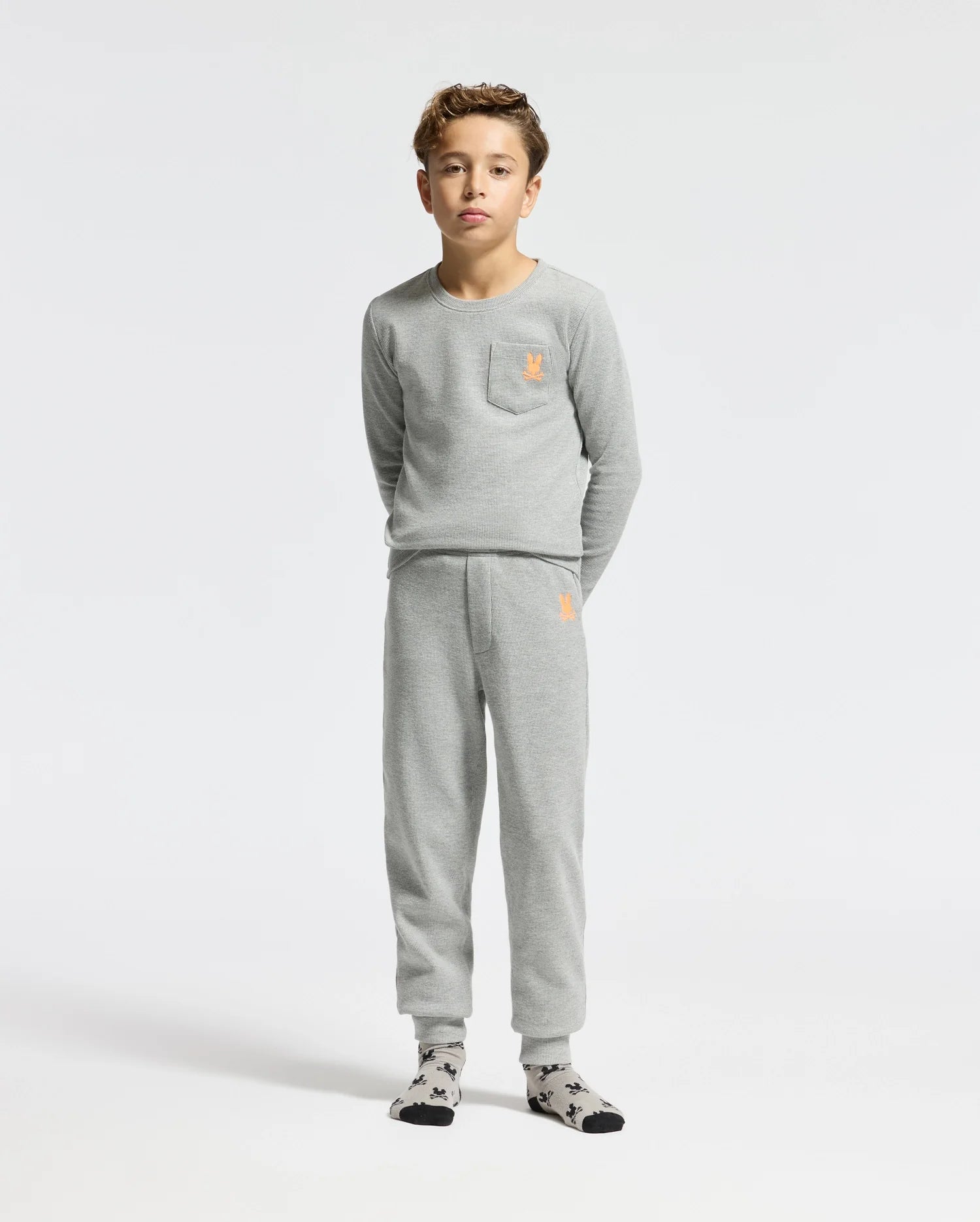A young boy stands wearing the Psycho Bunny KIDS VAL WAFFLE LONG SLEEVE TEE, which comes in a gray color with a matching top and bottom set. The pajamas feature an embroidered orange emblem on both the chest and pants. He is also wearing socks with a black and white pattern against a plain white background.