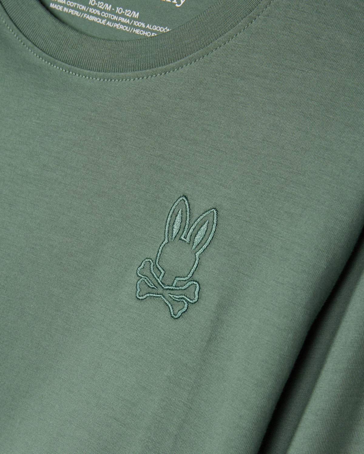 The KIDS ALBERTON LONG SLEEVE TEE (Model: B0T867D200) from Psycho Bunny is a green shirt made from soft Pima cotton, featuring a stitched emblem of rabbit ears and crossbones. Ideal for layering, the fabric label on the inside of the collar is partially visible.