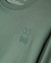The KIDS ALBERTON LONG SLEEVE TEE (Model: B0T867D200) from Psycho Bunny is a green shirt made from soft Pima cotton, featuring a stitched emblem of rabbit ears and crossbones. Ideal for layering, the fabric label on the inside of the collar is partially visible.