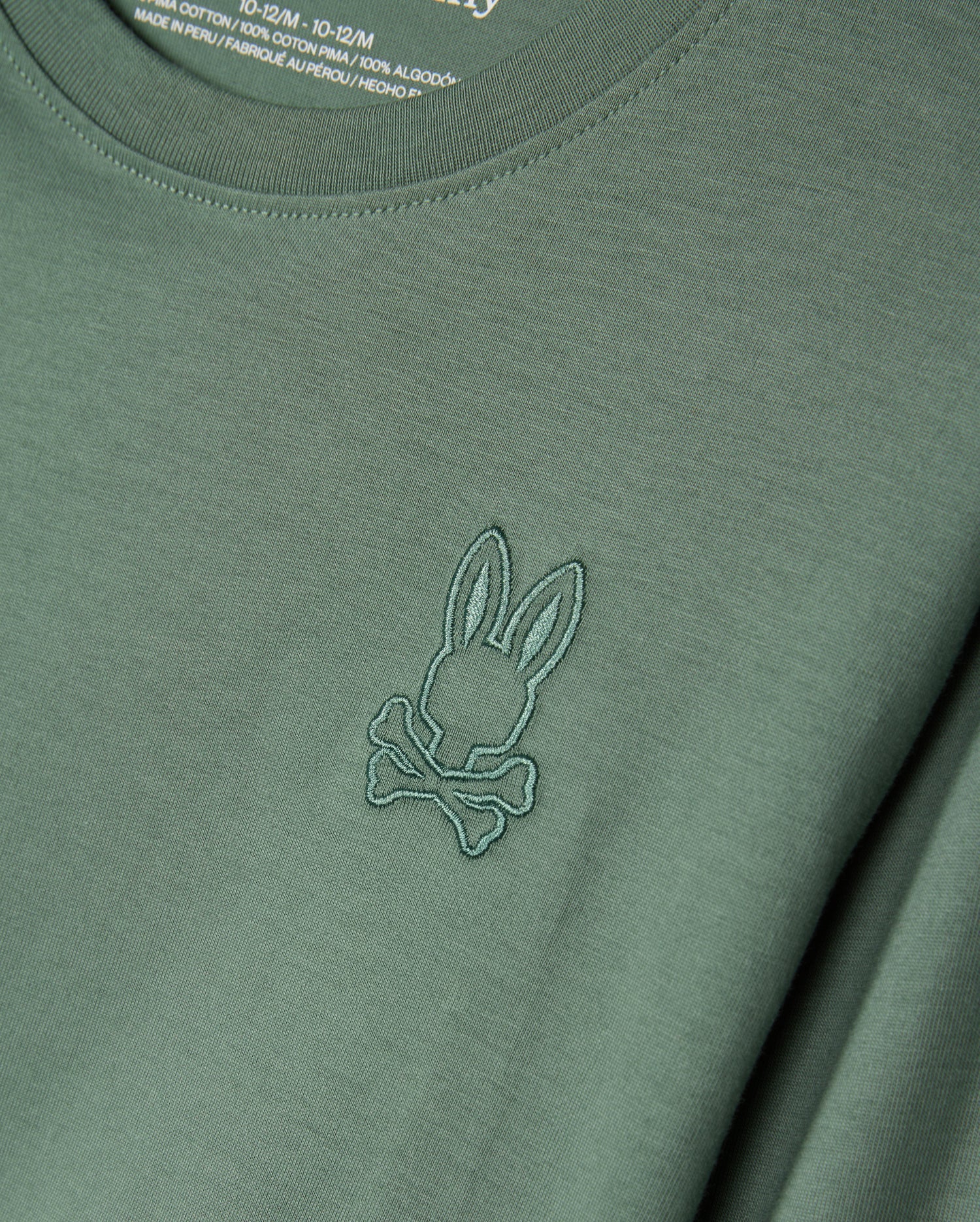 The KIDS ALBERTON LONG SLEEVE TEE (Model: B0T867D200) from Psycho Bunny is a green shirt made from soft Pima cotton, featuring a stitched emblem of rabbit ears and crossbones. Ideal for layering, the fabric label on the inside of the collar is partially visible.