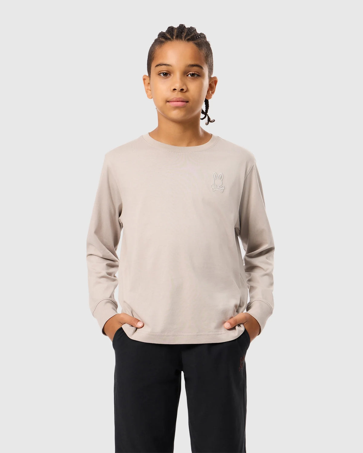 A person with braided hair stands with hands in pockets, wearing a light beige Psycho Bunny Kids Alberton Long Sleeve Tee (B0T867D200) and black pants, against a plain gray background.