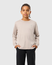 A person with braided hair stands with hands in pockets, wearing a light beige Psycho Bunny Kids Alberton Long Sleeve Tee (B0T867D200) and black pants, against a plain gray background.