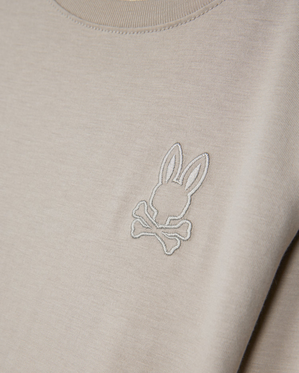 Introducing the Psycho Bunny KIDS ALBERTON LONG SLEEVE TEE - B0T867D200: A close-up showcases this beige, long sleeve tee crafted from soft Pima cotton. It features an embroidered logo of a stylized bunny head above crossed bones, delivering a playful twist on the classic skull and crossbones motif, all while ensuring comfort and style with its regular fit.