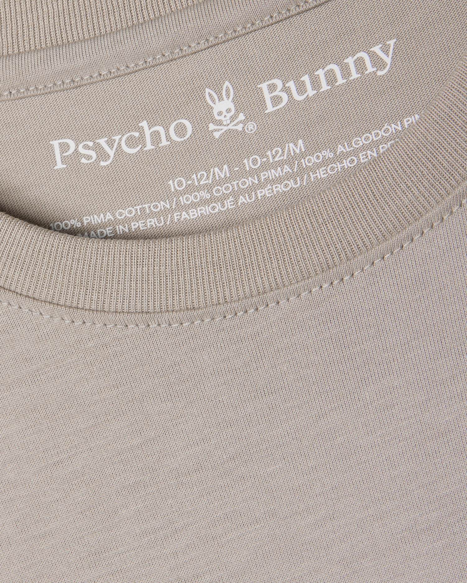 Close-up of a beige KIDS ALBERTON LONG SLEEVE TEE - B0T867D200 by Psycho Bunny, highlighting the brand logo and size details (10-12/M) inside the neckline. This regular fit tee is made from 100% Pima cotton for exceptional softness and quality, crafted in Peru.