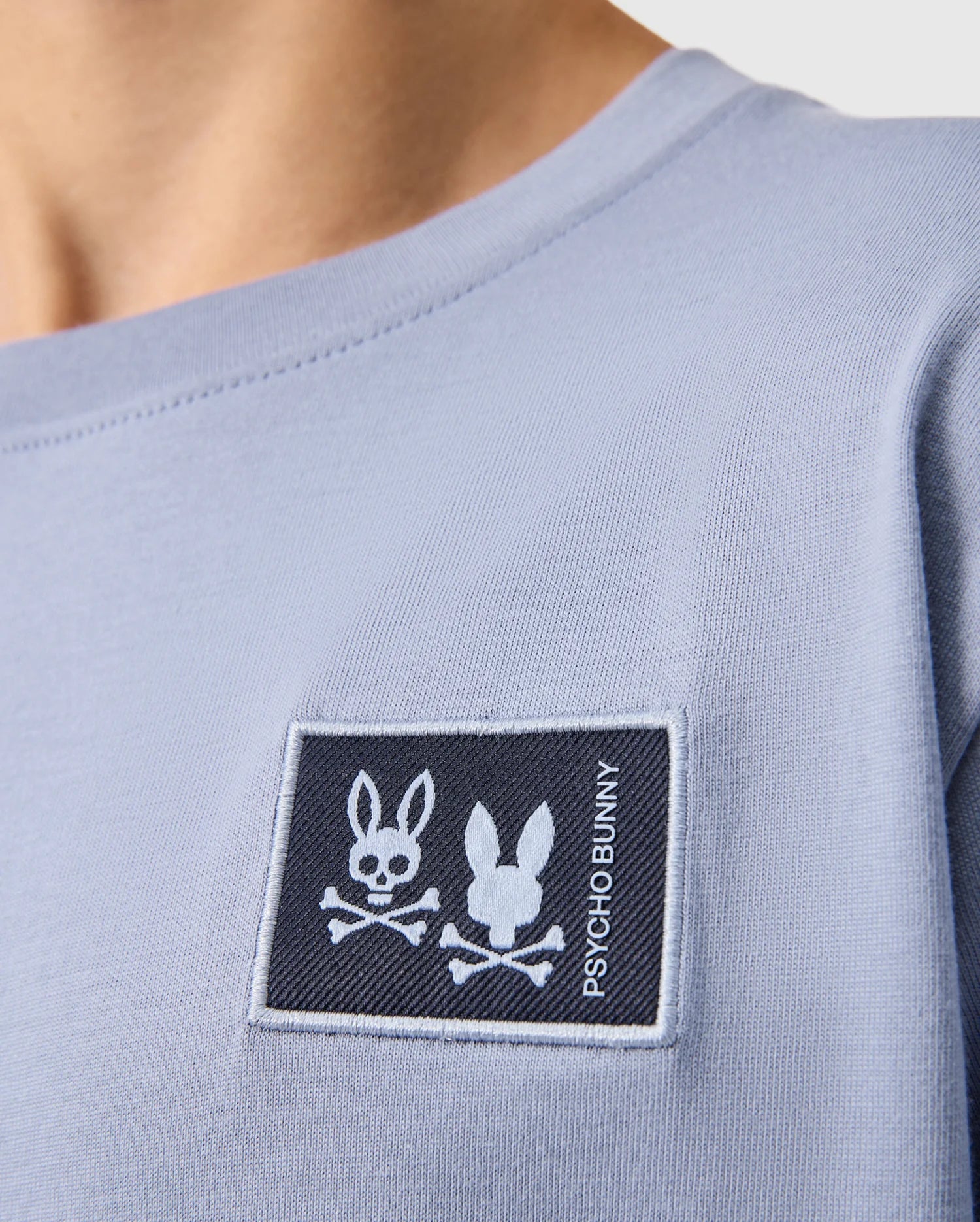 Close-up of someone wearing a Psycho Bunny Kids Monroe Long Sleeve Tee in light blue, featuring a dark blue rectangular chest patch with a bunny skull and crossbones design and "PSYCHO BUNNY," crafted from pima cotton.