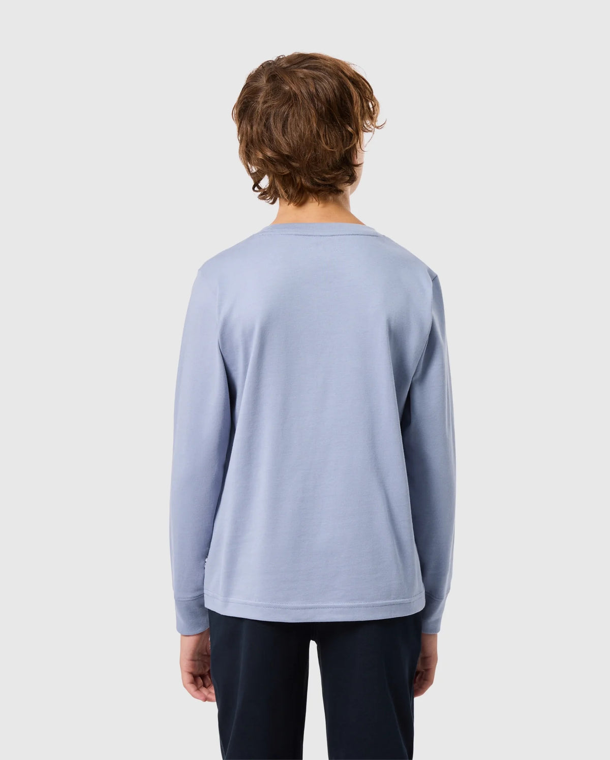 A person with medium-length brown hair stands facing away, wearing a Psycho Bunny KIDS MONROE LONG SLEEVE TEE in light blue and dark pants. The background is plain gray.