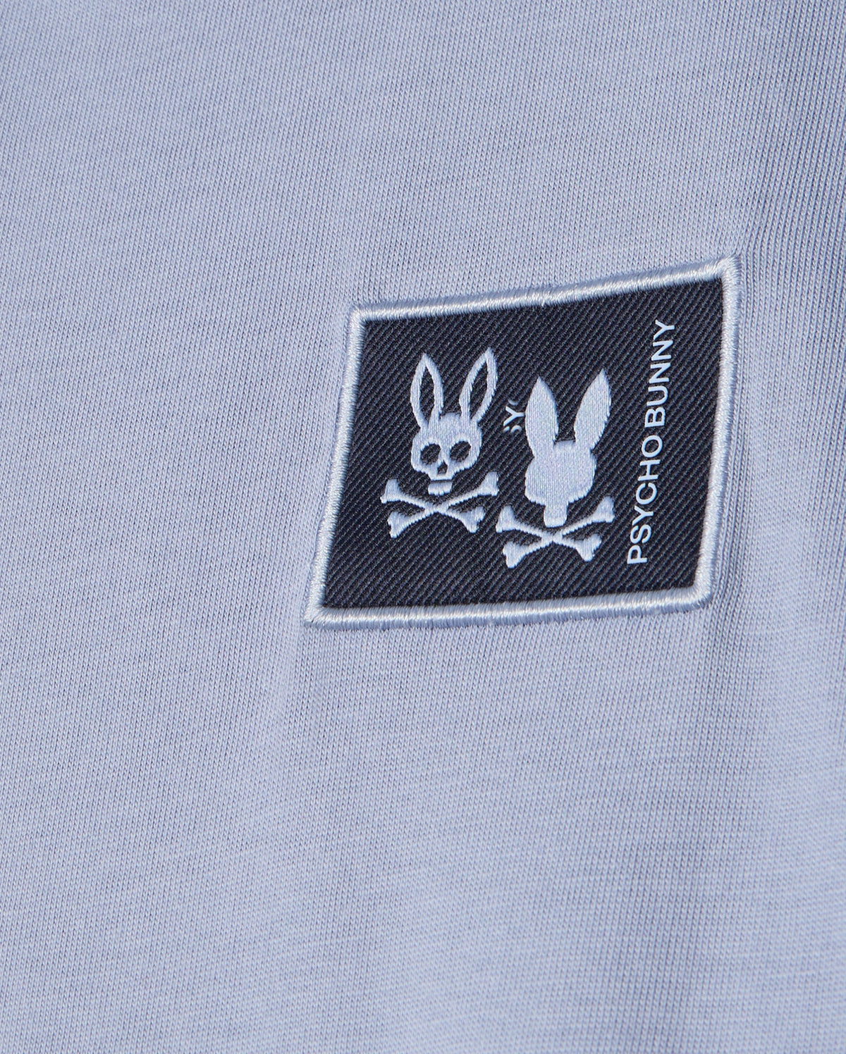 A close-up of the light blue Kids Monroe long sleeve tee in a regular fit features a black patch with Psycho Bunny's logo: two bunny heads with crossed bones—one upright, the other flipped. The brand name is written vertically on the patch for a versatile, cozy style.