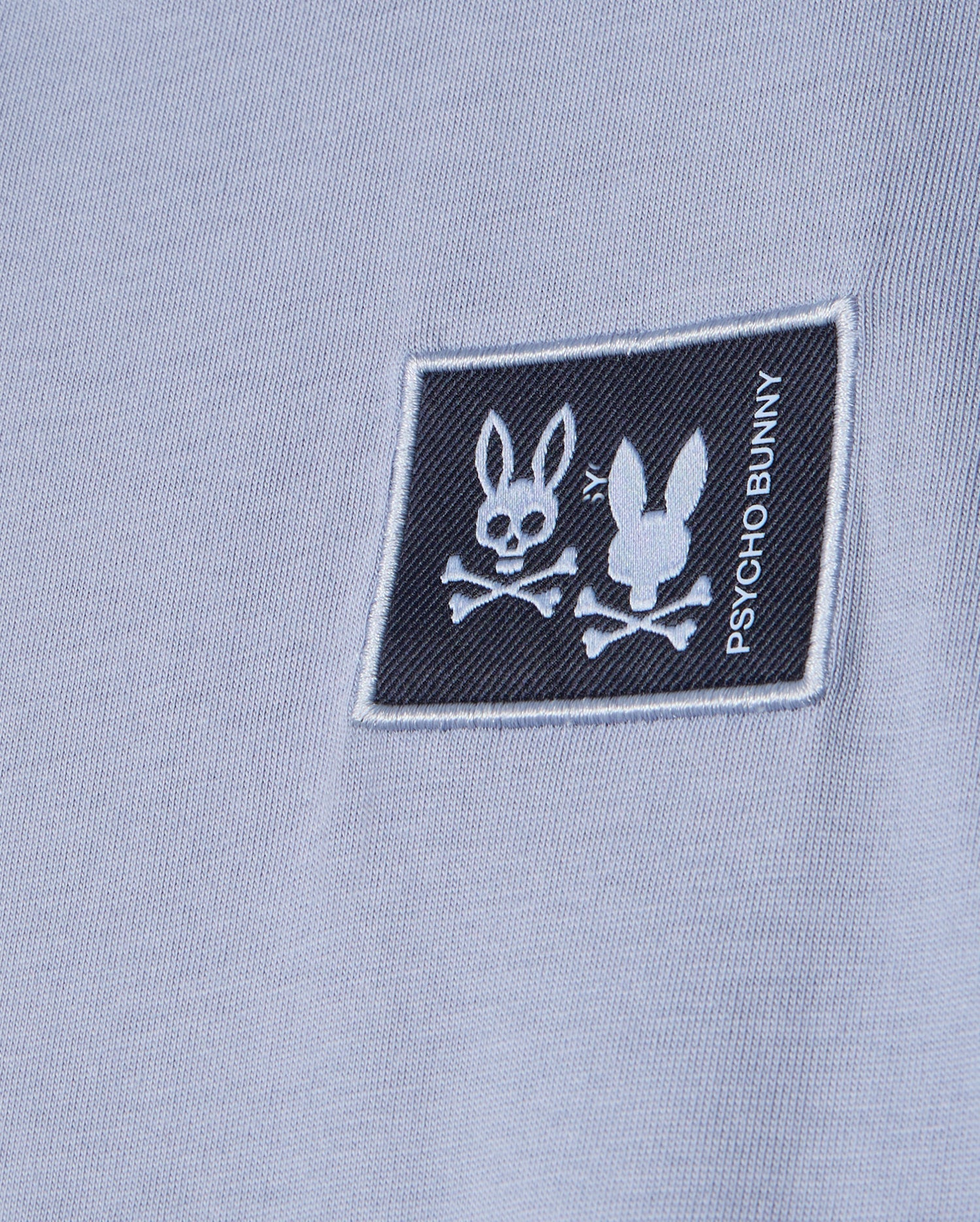 A close-up of the light blue Kids Monroe long sleeve tee in a regular fit features a black patch with Psycho Bunny's logo: two bunny heads with crossed bones—one upright, the other flipped. The brand name is written vertically on the patch for a versatile, cozy style.