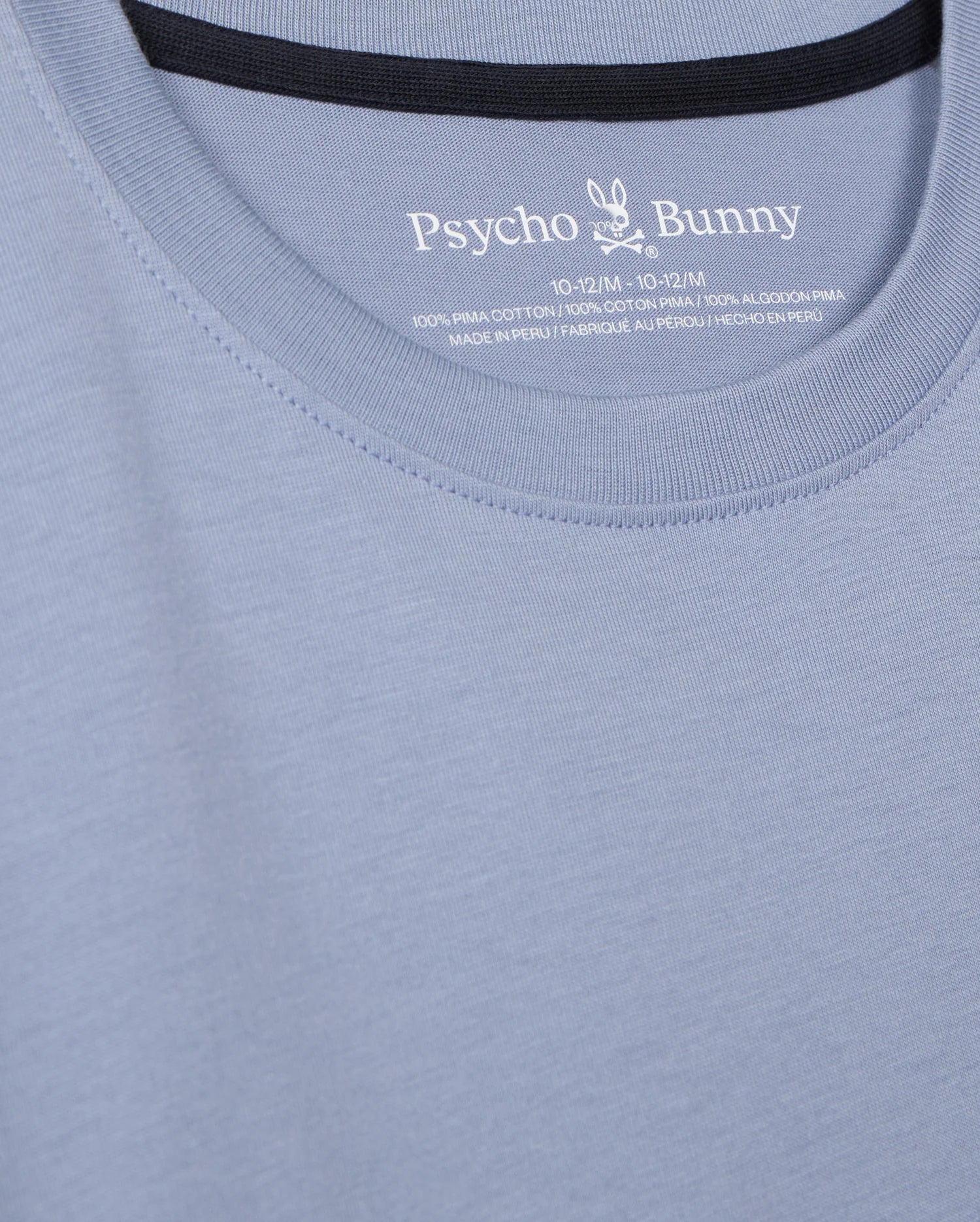 Close-up of the KIDS MONROE LONG SLEEVE TEE by Psycho Bunny, in light blue with logo and brand name on the inside collar. Made from pima cotton for a soft feel, it features cozy stitching details and texture, offering a regular fit that embodies versatile style. Model: B0T975D200.
