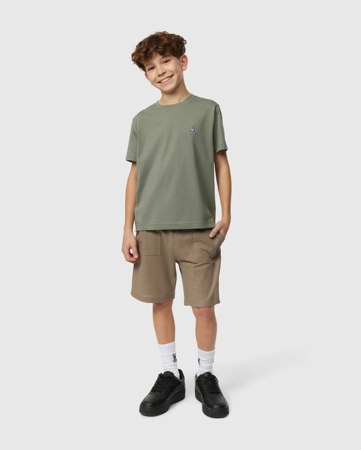 A smiling young boy with curly hair stands against a plain background. He is wearing a green KIDS CLASSIC CREW NECK TEE (B0U014B200) from Psycho Bunny, made from 100% Pima cotton as part of his back-to-school outfit, along with tan shorts, white socks, and black sneakers. One hand is in his pocket while the other hangs by his side.
