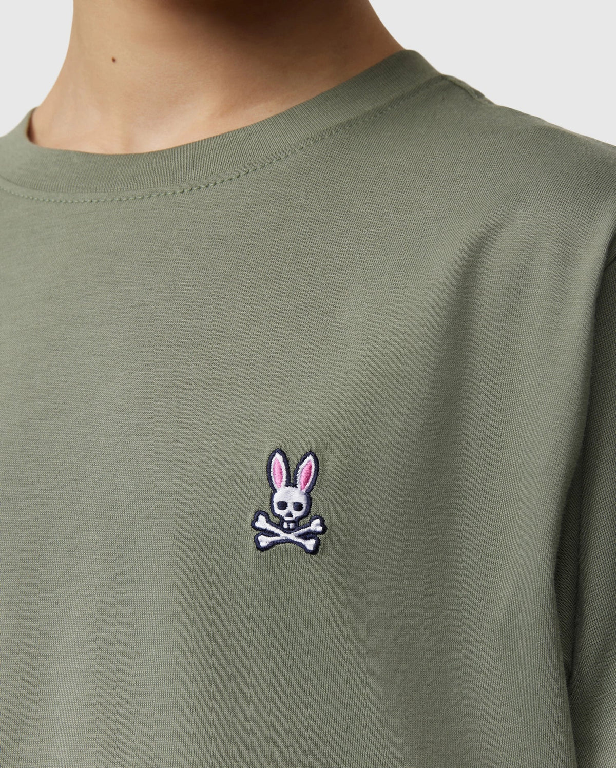 Close-up of a person wearing the KIDS CLASSIC CREW NECK TEE - B0U014B200 by Psycho Bunny in olive green. Crafted from 100% Pima cotton, the shirt features a small embroidered logo on the chest, showcasing a skull and crossbones with bunny ears in pink. Perfect for back-to-school outfits, it’s one of those kids' classic crew neck tee shirts they'll love.